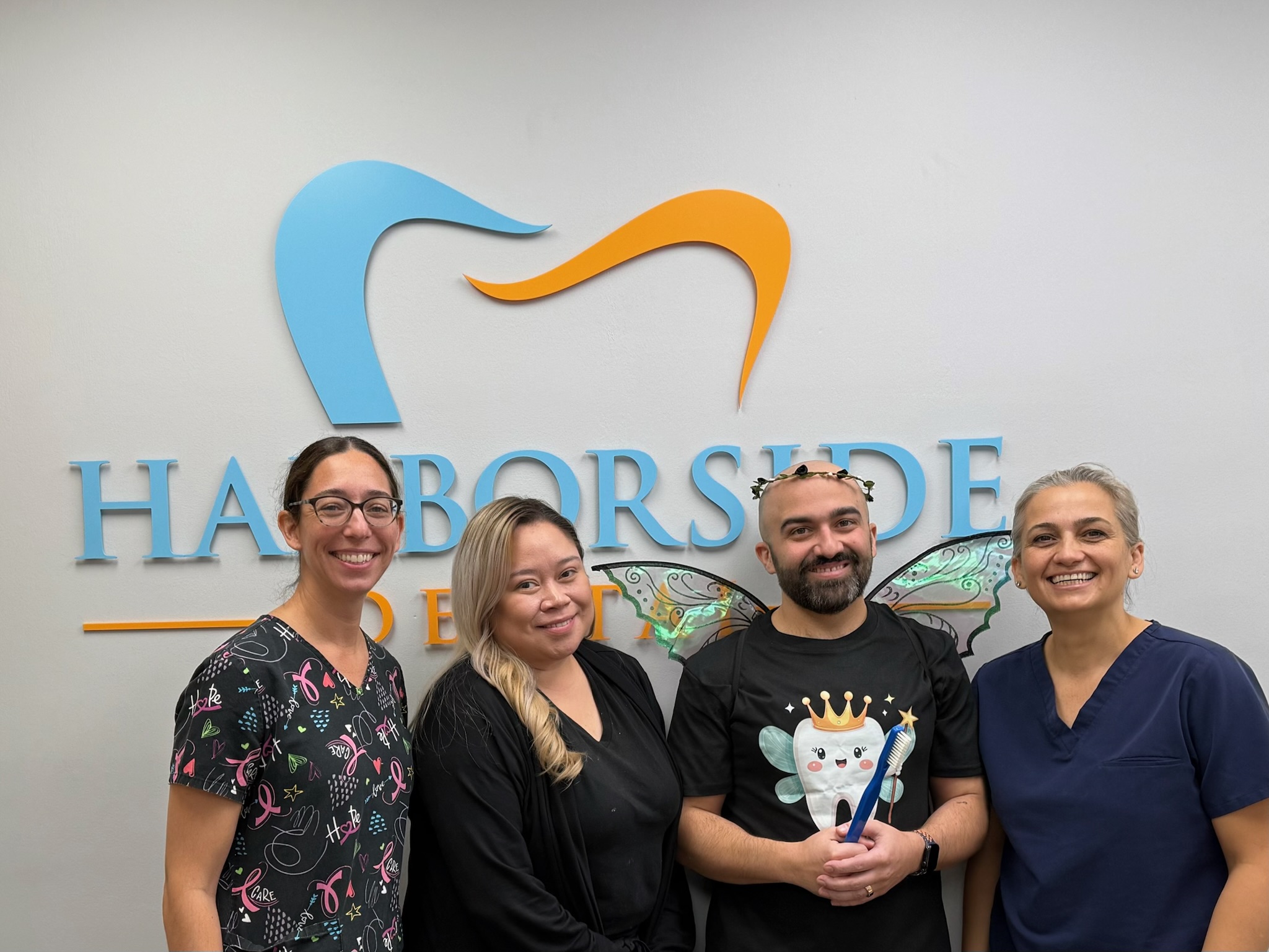 Jahan "Dr. Jay" Monirian and his team of smiling dental professionals posing together in front of a colorful dental office sign that reads 'Harborside Dental of Winthrop.'